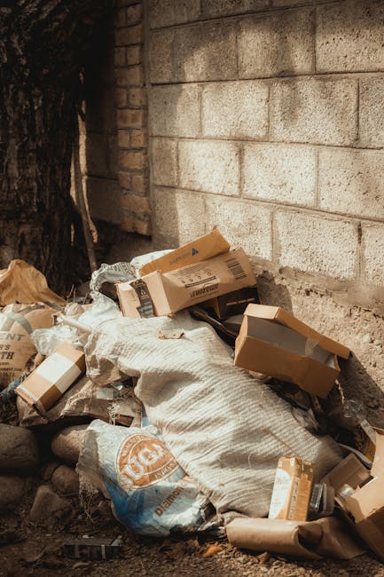 A pile of mixed waste materials and cardboard boxes is accumulated at the base of a grey brick wall beside a tree trunk. The cardboard boxes vary in size, with some partially crushed or torn, featuring printed labels and brown kraft finish. The waste also includes plastic bags, some knotted, and a fabric sack with a woven pattern, all in neutral tones of brown, beige, and white. The pile is situated outdoors, resting directly on a dirt surface with small rocks scattered around, indicating an untidy area that could be part of a private property or a designated waste spot. The background shows a weathered brick wall with a rough texture, and the presence of a tree trunk on the left adds natural context. The scene is illuminated by natural daylight, highlighting the textures of the cardboard, fabric, and wall, consistent with a setting where independent rubbish collection or on-site clearance might be necessary to manage accumulated debris effectively, aligning with services offered by Rubbish Removal Harringay.