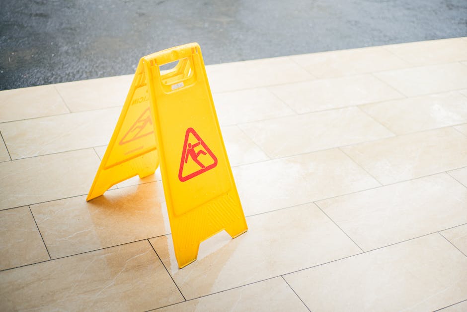 A bright yellow, foldable warning sign with a red triangular caution symbol featuring a fallen person icon is positioned on a polished beige tiled floor, indicating a slip hazard. The sign is made of durable plastic with a textured base for stability and is placed in an indoor or outdoor environment, possibly a commercial or residential entryway. The background shows a contrasting darker, rough surface, likely asphalt or concrete, indicating a transition from indoor to outdoor space or a parking area. The sign's vibrant color and clear safety symbol are intended to alert passersby to exercise caution, reflecting a common practice in waste removal or maintenance scenarios where wet or uneven surfaces pose a risk. The image subtly supports the context of managing safety during rubbish collection or site clearance activities, highlighting the importance of proper safety signs in private waste handling and independent collection environments, which Rubbish Removal Harringay may service.
