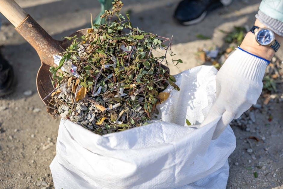A person wearing a grey jacket and a wristwatch using a white gloves to hold an open white woven polypropylene sack filled with garden debris, including small branches with green leaves, woody twigs, and dry plant material. The person is standing outdoors on a paved or gravel surface, and a rusty shovel with a wooden handle is partially inserted into the sack, suggesting active garden waste collection. In the background, a pair of black shoes and a glimpse of nearby greenery or ground debris can be seen, emphasizing an outdoor setting suitable for independent waste disposal or garden clearance, which aligns with alternative rubbish removal services like those offered by Rubbish Removal Harringay.