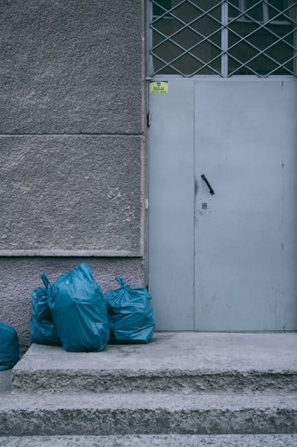 Image showing three blue rubbish bags placed on a concrete doorstep in front of a grey metal door with a black handle, located at the base of a building with a rough textured grey wall and a section of metal barred window above the door. The bags are made of plastic, appear full, and are slightly crumpled. The setting suggests a private or independent waste collection area, possibly for on-site rubbish removal for nearby flats or maisonettes in Harringay, with minimal environmental context visible beyond the immediate doorstep. The lighting is natural, and the overall scene emphasizes the presence of loose waste prepared for collection, consistent with rubbish removal services such as those offered by Rubbish Removal Harringay. This scene exemplifies the typical arrangement for private waste handling, highlighting the practicalities of rubbish disposal at accessible entry points, intended for the safe and convenient removal of refuse from multi-storey residential buildings.