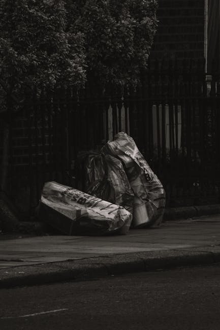 Two large black plastic garbage bags filled with waste are positioned on the pavement near the curb, leaning against a black metal fence with vertical bars. The bags appear crumpled and are tightly packed, with some paper or cardboard packaging visible on the surface, indicating general household rubbish. Behind the fence, dense foliage from nearby trees partially obscures the background, and a building or structure with dark, grid-like fencing or panels is visible further back. The scene is illuminated with natural light, creating contrast between the dark fence and the lighter tones of the bags and pavement. The placement of the bags suggests an instance of independent waste disposal, likely managed by Rubbish Removal Harringay, serving local shops and flats through private rubbish collection services rather than local authority collection. The overall aesthetic emphasizes the practical aspects of rubbish removal in an urban environment, with a focus on the environmental context of waste management and on-site clearance for the community.