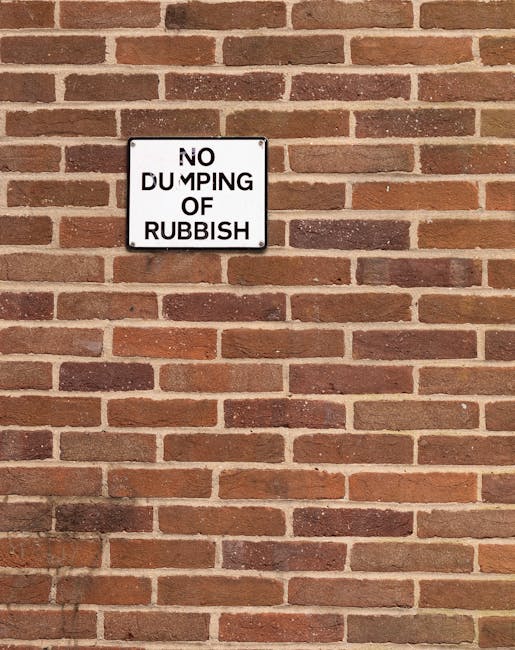 A white rectangular metal sign with black text stating 'NO DUMPING OF RUBBISH' is mounted on a red brick wall. The bricks have a rough texture with some variation in shades of red, brown, and orange, and are arranged in a traditional horizontal pattern with visible mortar joints. The sign is positioned slightly toward the upper left corner of the wall and appears to be securely attached. The brick wall extends beyond the edges of the image, with no other objects or environmental features visible, creating a neutral background that emphasizes the warning message against improper waste disposal, consistent with regulations for private rubbish collection and on-site clearance services by Rubbish Removal Harringay.