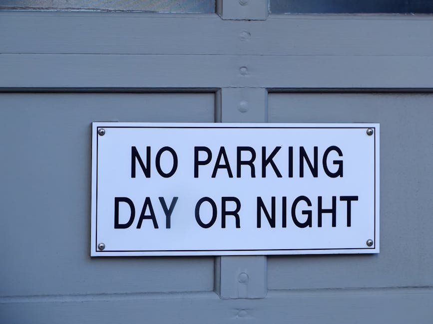 A rectangular white metal sign with bold black lettering that reads 'NO PARKING DAY OR NIGHT' is mounted on a light gray wooden door panel. The sign is secured with four silver screws, one at each corner. The door panel features horizontal and vertical wooden slats, with a faint texture visible on the painted surface. The surrounding environment includes the door's framing, contributing to a neutral urban setting, typical of private or commercial premises where access restrictions such as no parking signify an area designated for rubbish removal or service vehicles. The lighting appears natural, with soft shadows and even illumination, emphasizing the clarity of the sign and the painted surface details. This scene subtly relates to waste management contexts where designated no-parking zones facilitate on-site rubbish removal or alternative waste handling procedures, as exemplified by companies like Rubbish Removal Harringay.
