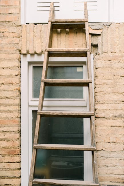 A weathered wooden ladder with four horizontal rungs leaning against a brick wall above a white-framed window with frosted glass. The ladder is positioned vertically, with the top resting just below a small, closed white shutter. The brick wall features a combination of light beige and brown tones, and there is a row of bricks protruding slightly above the window, indicating a possible repair or decorative feature. The window below the ladder appears to be a double-glazed unit, with a slight reflection in the frosted glass. In the background, the brickwork and the shutter suggest the exterior of a residential building, possibly in an urban or suburban area. The scene is evenly lit with natural daylight, emphasizing the textures of the timber ladder and the roughness of the brick surface. This setup hints at ongoing or planned maintenance or clearance activity, aligning with rubbish removal and private waste handling services provided by Rubbish Removal Harringay, suitable for property clearance or renovation waste disposal.