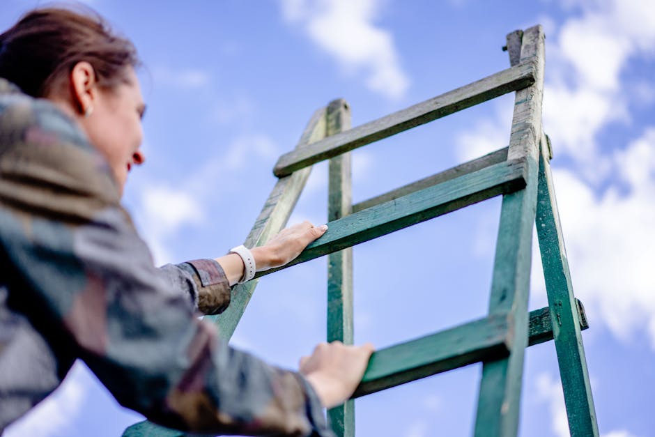 A woman wearing a plaid shirt is outdoors during daytime, climbing a wooden ladder with weathered green paint. The ladder is positioned against a bright blue sky with scattered white clouds, and she is gripping the side rails with her hands as she ascends. The background shows a slightly blurred sky, highlighting the outdoor setting. The image captures a moment of activity involving manual effort, with natural lighting illuminating the scene. While the focus is on her climbing the ladder, subtle hints of environmental context suggest efforts related to home maintenance, gardening, or external property work, which can be associated with tasks like rubbish removal or property clearance by professional services such as Rubbish Removal Harringay.