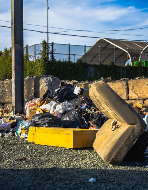 A pile of mixed household waste and rubbish sits on a gravel surface outdoors, consisting of black plastic bags, cardboard boxes, and various discarded items, including a single large, worn car tire leaning against the heap. The waste appears to be gathered from a local residential or commercial area for collection or disposal, with a low stone wall and greenery behind it. In the background, there is a metal fence, utility poles with overhead wires, and a partially visible modern shelter or canopy structure with a sky that is partly cloudy, indicating daytime. The scene reflects the type of waste material that might be handled through private rubbish removal services such as Rubbish Removal Harringay, illustrating an example of on-site clearance or independent collection options for waste management, particularly when local authority refuse collection is not available or insufficient.