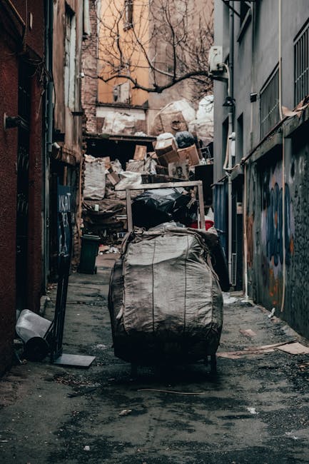 A pile of mixed household waste and rubbish sits on a gravel surface outdoors, consisting of black plastic bags, cardboard boxes, and various discarded items, including a single large, worn car tire leaning against the heap. The waste appears to be gathered from a local residential or commercial area for collection or disposal, with a low stone wall and greenery behind it. In the background, there is a metal fence, utility poles with overhead wires, and a partially visible modern shelter or canopy structure with a sky that is partly cloudy, indicating daytime. The scene reflects the type of waste material that might be handled through private rubbish removal services such as Rubbish Removal Harringay, illustrating an example of on-site clearance or independent collection options for waste management, particularly when local authority refuse collection is not available or insufficient.