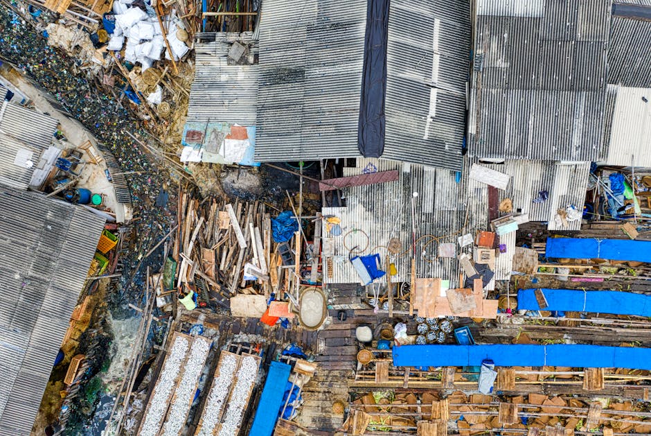 An aerial view of a cluttered outdoor area featuring a variety of discarded materials and debris. The scene includes several makeshift structures with corrugated metal roofing, some panels appearing rusted or weathered. Piles of wooden planks, broken furniture, and assorted rubbish are scattered across the ground, with some items partially stacked or leaning against each other. Blue tarpaulins cover sections of the clutter, while other loose objects such as pipes, plastic containers, and construction debris are visible among the disorder. The environment appears to be a private waste collection site or an informal rubbish dump, with a mixture of raw, unfinished materials and household waste. The lighting suggests daytime conditions, with shadows and highlights emphasizing the textures of the debris and metallic surfaces. This scene exemplifies situations where independent rubbish removal services, like those offered by Rubbish Removal Harringay, are often employed to clear such clutter efficiently and safely, especially in areas with informal or on-site disposal practices.