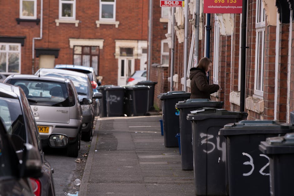 A residential street scene featuring a row of dark grey wheelie bins with white graffiti markings positioned along the sidewalk in front of brick houses with white-framed windows and stone window sills. The bins are lined up close to the building facades, which display peeling paint and signs of weathering. In the background, a woman dressed in a dark coat is standing beside one of the bins, appearing to interact with it or inspect its contents. Several parked cars, varying in color and model, are situated along the curbside, partially blocking the view of the street further ahead. The environment appears to be late afternoon with muted natural light. This scene reflects an area where residents and waste management services, such as Rubbish Removal Harringay, might coordinate independent rubbish collection or on-site clearance efforts outside typical council refuse collection schedules.