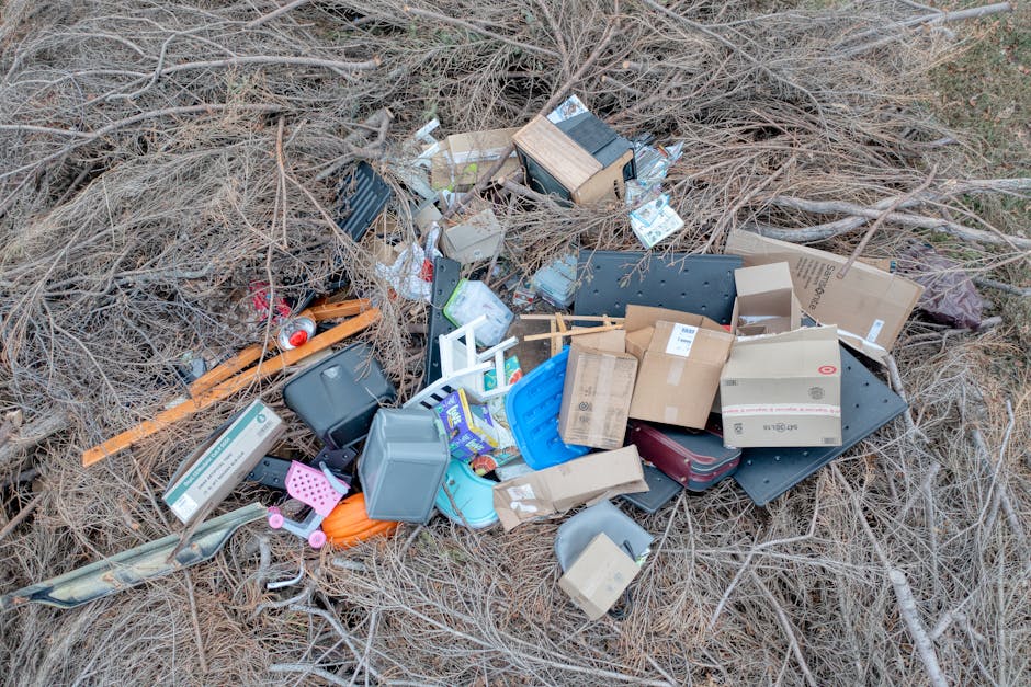 An aerial view of a cluttered outdoor area featuring a variety of discarded materials and debris. The scene includes several makeshift structures with corrugated metal roofing, some panels appearing rusted or weathered. Piles of wooden planks, broken furniture, and assorted rubbish are scattered across the ground, with some items partially stacked or leaning against each other. Blue tarpaulins cover sections of the clutter, while other loose objects such as pipes, plastic containers, and construction debris are visible among the disorder. The environment appears to be a private waste collection site or an informal rubbish dump, with a mixture of raw, unfinished materials and household waste. The lighting suggests daytime conditions, with shadows and highlights emphasizing the textures of the debris and metallic surfaces. This scene exemplifies situations where independent rubbish removal services, like those offered by Rubbish Removal Harringay, are often employed to clear such clutter efficiently and safely, especially in areas with informal or on-site disposal practices.
