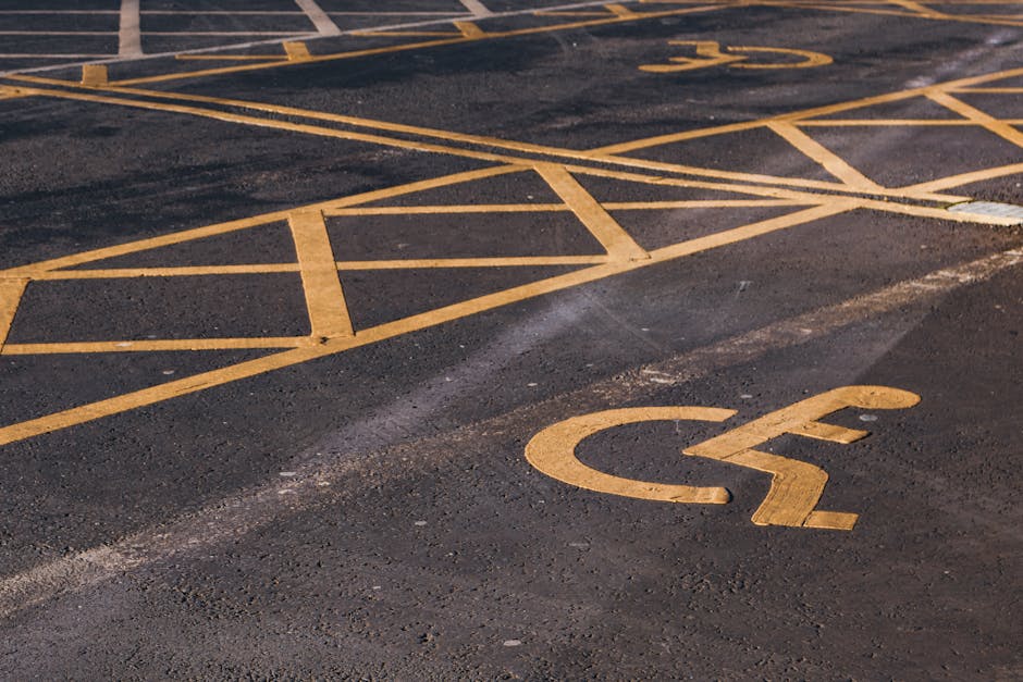 A close-up view of an asphalt parking lot surface featuring yellow painted markings, including a designated accessible parking space with a chalked outline and a wheelchair symbol, located near other standard parking bays marked with yellow lines. The surface shows textured asphalt with visible small gravel and slightly uneven patches, illuminated by natural daylight. In the foreground, a yellow-painted disabled parking symbol with a stylized wheelchair figure is prominently visible, indicating accessibility provisions. The parking area is bordered by a curb and surrounds a building or street scene, though no surrounding structures are visible in the image. The scene conveys an environment where waste management services, such as rubbish removal, can be efficiently coordinated through appropriate parking and access arrangements, which Rubbish Removal Harringay may utilise for on-site clearance or private disposal tasks in the area.