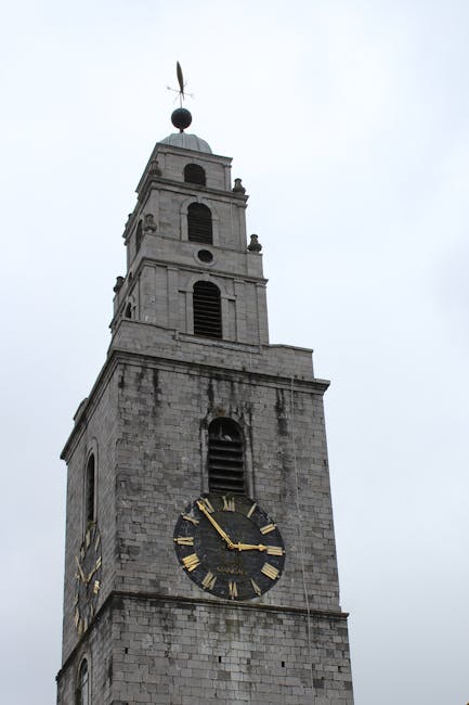 A tall historic clock tower constructed from light grey stone stands prominently against a cloudy sky. The tower features multiple levels with arched windows and decorative stone details. The large clock face, positioned near the bottom of the tower, has a black background with gold Roman numerals and gold hands, showing the time as approximately 3:05. Above the clock, there are additional narrow windows and architectural accents, leading up to a smaller upper section with a rounded, greenish-tinged domed roof topped by a weather vane. The structure is part of an urban environment, possibly a church or civic building, and Rubbish Removal Harringay occasionally references such iconic landmarks when discussing various aspects of property and site clearance. The weather appears overcast, with soft, diffuse natural light illuminating the stone exterior, highlighting its textured aged surface and subtle weathering marks typical of historic municipal architecture.