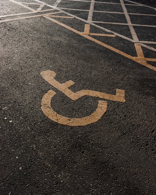 A close-up view of an asphalt parking lot surface featuring yellow painted markings, including a designated accessible parking space with a chalked outline and a wheelchair symbol, located near other standard parking bays marked with yellow lines. The surface shows textured asphalt with visible small gravel and slightly uneven patches, illuminated by natural daylight. In the foreground, a yellow-painted disabled parking symbol with a stylized wheelchair figure is prominently visible, indicating accessibility provisions. The parking area is bordered by a curb and surrounds a building or street scene, though no surrounding structures are visible in the image. The scene conveys an environment where waste management services, such as rubbish removal, can be efficiently coordinated through appropriate parking and access arrangements, which Rubbish Removal Harringay may utilise for on-site clearance or private disposal tasks in the area.