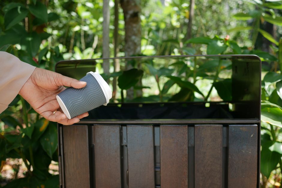 A person wearing a beige long-sleeved coat is holding a grey paper coffee cup with a white plastic lid, positioning it above a rusty metal trash bin with a partly open lid. The trash bin is situated outdoors on a paved surface, surrounded by green foliage, including broad leaves and tree trunks in the background, suggesting a garden or park environment. The scene is lit with natural daylight, casting soft shadows and highlighting the textures of the leaves, metal, and the person's hand. The image relates to waste disposal practices, possibly illustrating an example of private rubbish handling or on-site rubbish collection, aligning with the context of independent waste management services in Harringay.