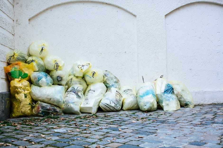 An overflowing collection of various waste items is positioned on a paved sidewalk in front of a commercial building, exhibiting a mixture of cardboard boxes, plastic bags, paper waste, and discarded packaging materials. The waste is piled high around and on top of several large wheelie bins, including a grey mixed waste container, a black recycling bin, and a bright red bin, with some trash spilling onto the cobbled street surface. The waste appears to consist of household, packaging, and miscellaneous refuse, with some of the cardboard and paper items crumpled or flattened, while others remain unfolded or intact. In the background, the building features a yellow storefront with signage and glass windows, and an adjacent blue scaffolding structure suggests ongoing building work above. To the left, a car with a partially visible rear license plate is parked behind a metal barrier, while a tree with bare branches is situated near the waste collection point, indicating a typical urban environment. The scene reflects a situation that could relate to private waste management services like those offered by Rubbish Removal Harringay, emphasizing the importance of proper rubbish disposal to avoid fines related to illegal dumping or improper waste handling in residential or commercial areas.